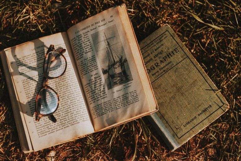 From above of old textbooks with illustration and eyewear on open page on dry grass in sunlight